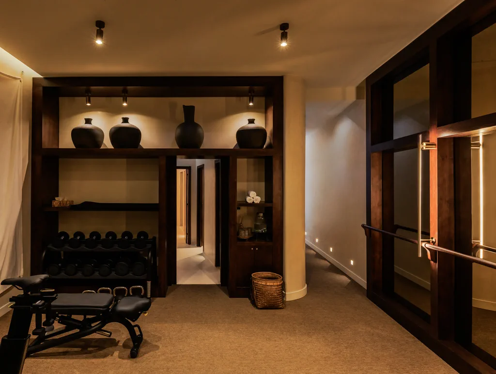 The gym in warm low light, dumbbells and a bench by dark wood shelves