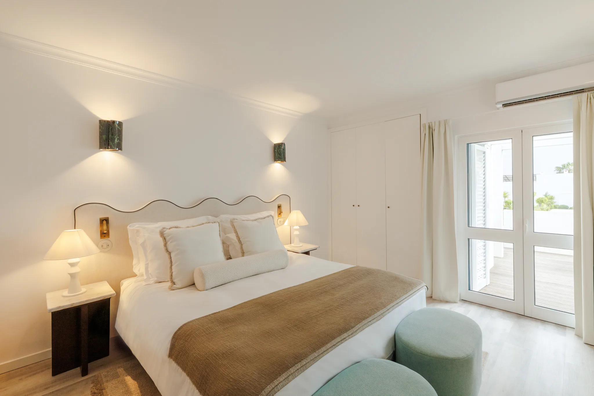 A bedroom with the bedside lamps lit and two sage poufs at the foot of the bed
